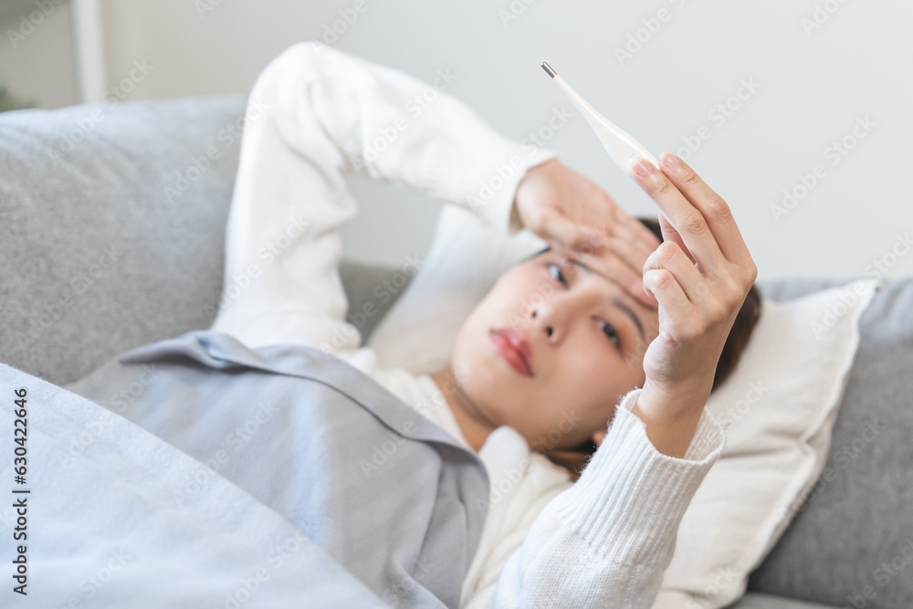 Sick, influenza asian young woman, girl have a fever, hand holding thermometer, touching forehead for check measure body temperature, illness while lying rest on sofa at home. Health care concept. Sick, influenza asian young woman, girl have a fever, hand holding thermometer, touching forehead for check measure body temperature, illness while lying rest on sofa at home. Health care concept.