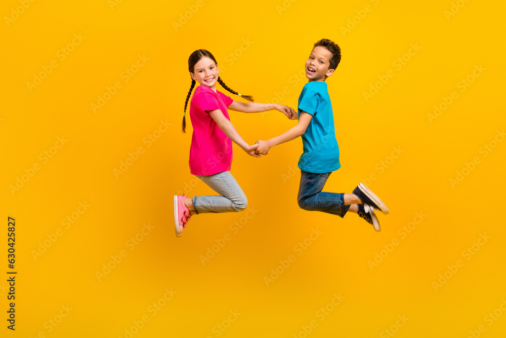 Full size profile portrait of two carefree cute kids jumping hold arms rejoice isolated on yellow color background