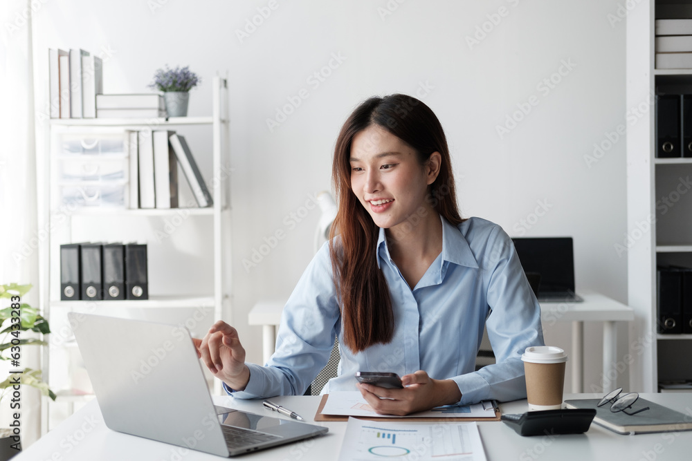 Beautiful Asian businesswoman working with laptop computer while holding smartphone in her hand.