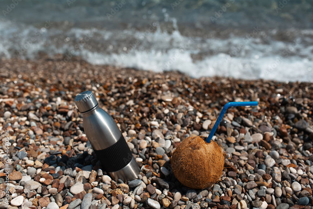 Close-up of steel thermo water bottle and coconut cocktail on the stone beach against sea waves. Close-up of steel thermo water bottle and coconut cocktail on the stone beach against sea waves.