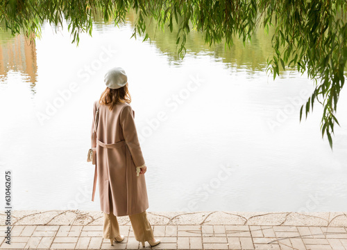 A young girl in a beige coat stands on the shore of a lake in a park. Close-up. Selective focus. Autumn landscape.