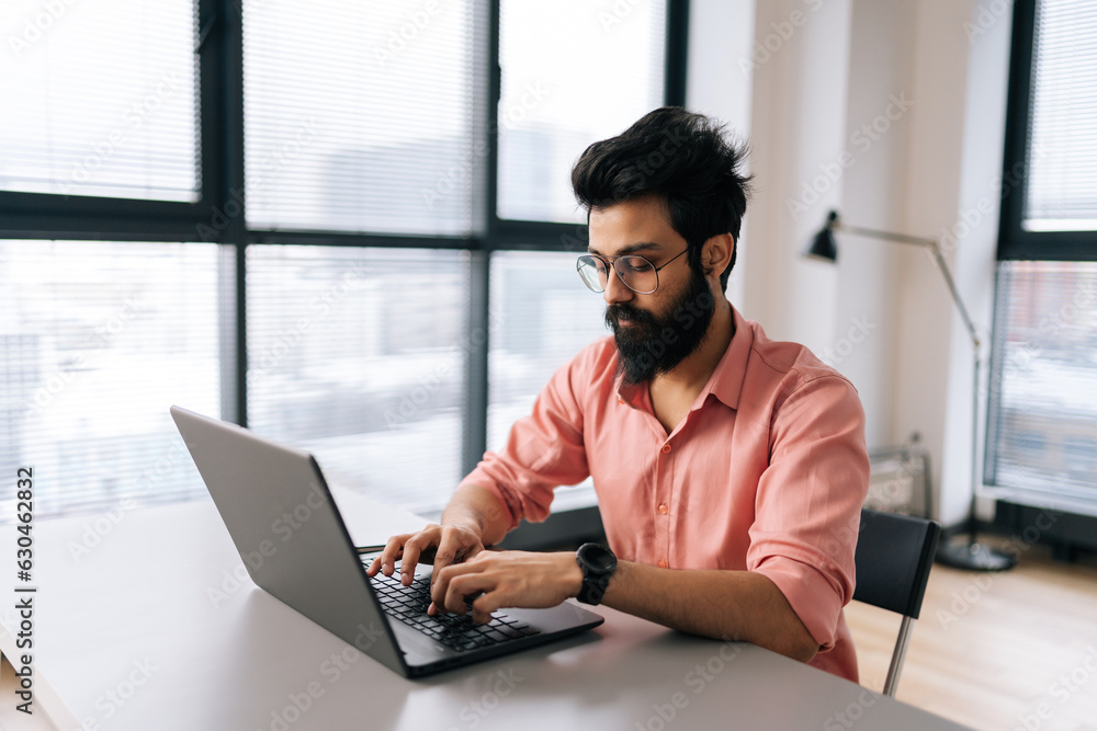 Indian designer male in stylish eyeglasses working on laptop sitting at desk in light coworking office room by window. Bearded business man wearing casual clothes looking at computer screen. Indian designer male in stylish eyeglasses working on laptop sitting at desk in light coworking office room by window. Bearded business man wearing casual clothes looking at computer screen.