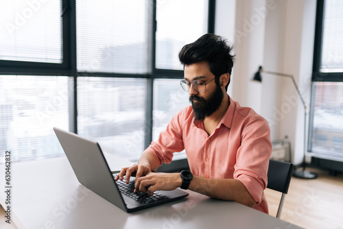 Indian designer male in stylish eyeglasses working on laptop sitting at desk in light coworking office room by window. Bearded business man wearing casual clothes looking at computer screen.