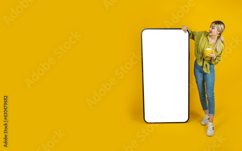 Full body young blonde girl stand near leaning big huge smartphone blank screen mobile cell phone mock up. Looking empty display. Application, recommend, offer, banner design concept. Copy space.