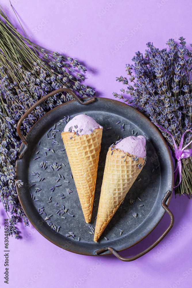 Two lavender ice creams in waffle cones on a tin tray and bouquets of dry lavender on a lilac background.