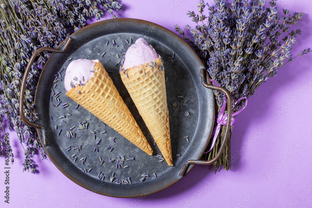 Two lavender ice creams in waffle cones on a tin tray and bouquets of dry lavender on a lilac background.