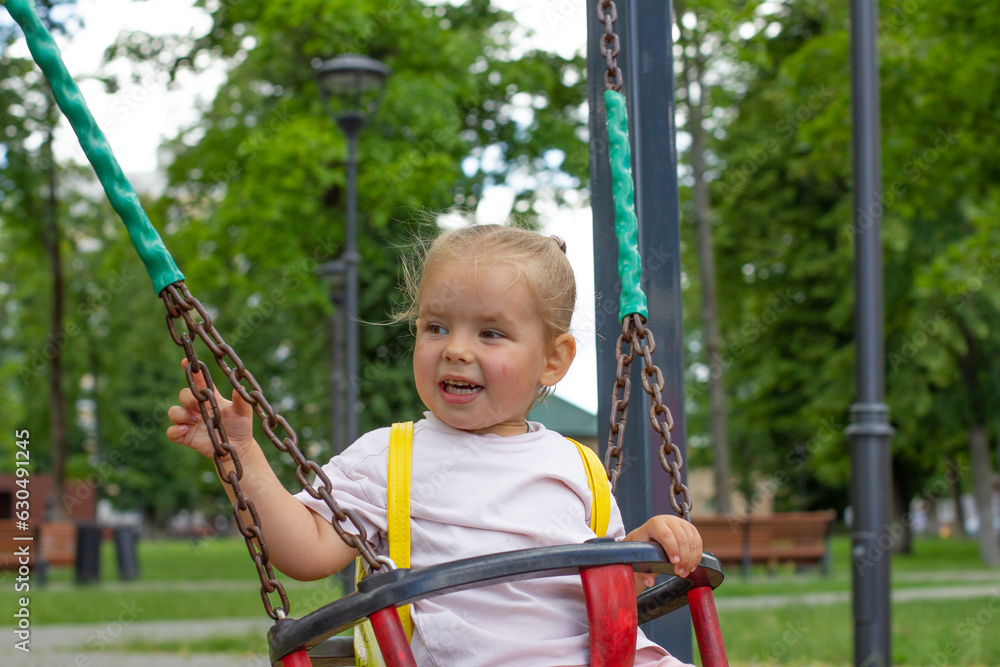 A cute little girl swings on a swing in the park in summer and laughs. The child has fun in the summer in the park swinging on a swing. Happy child concept. A cute little girl swings on a swing in the park in summer and laughs. The child has fun in the summer in the park swinging on a swing. Happy child concept.