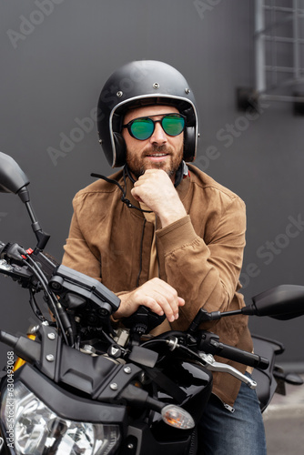 Smiling bearded man, biker wearing helmet, eyeglasses, leather jacket posing on motorcycle