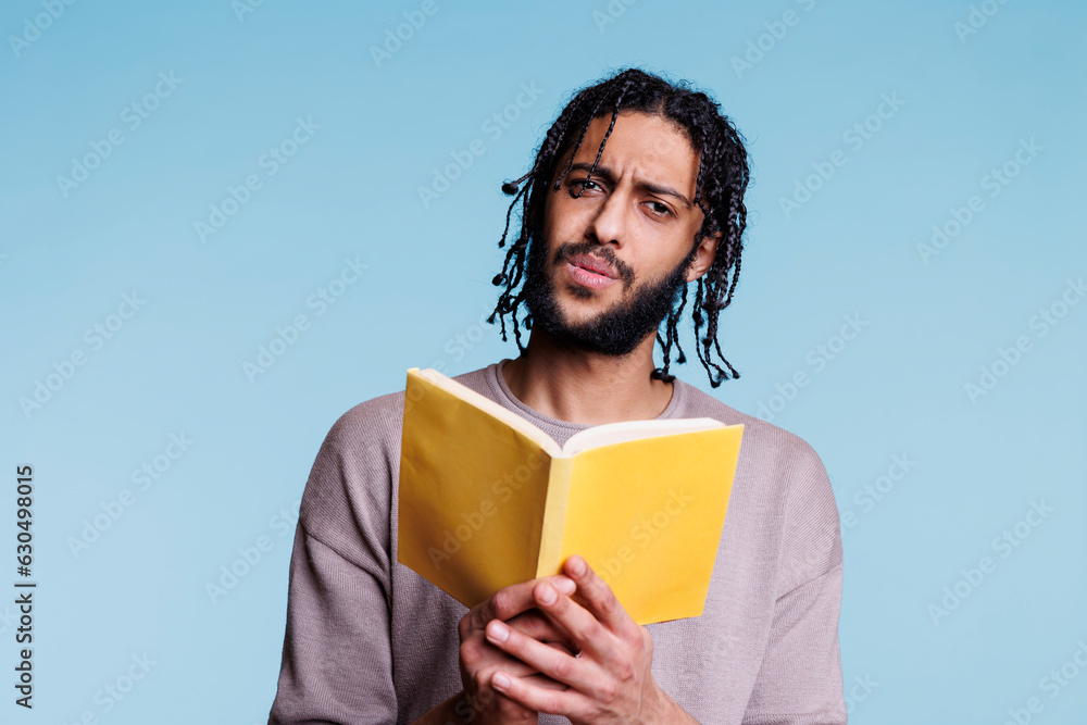 Confused arab man reading paperback book and looking at camera with puzzled facial expression. Young questioned person holding open softcover textbook and learning portrait