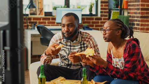 Happy life partners eating slices of pizza on couch, having fun together watching favorite movie on television. Young man and woman in relationship enjoying delivery food and alcohol.