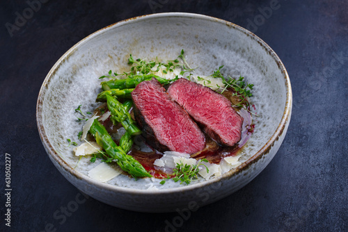 Gourmet barbecue dry aged angus beef steak with green asparagus and parmesan cheese in truffle mousse parfait and wild cherry relish cream served as close-up in a Nordic design bowl