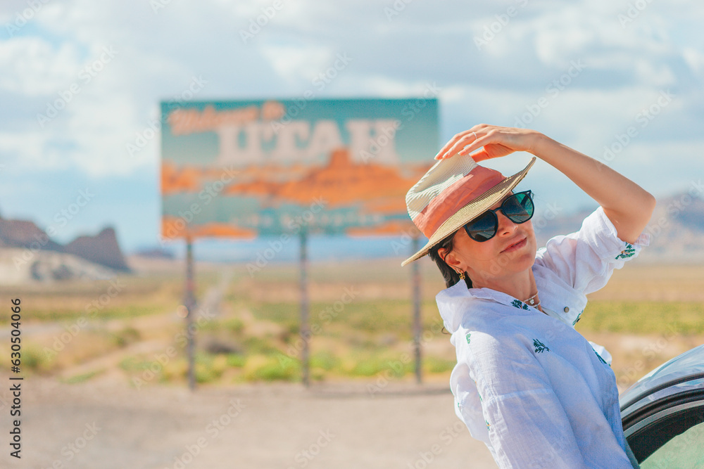 Beautiful woman on her trip by the car. Welcome to Utah road sign. Large welcome sign greets travels in Monument Valley, Utah