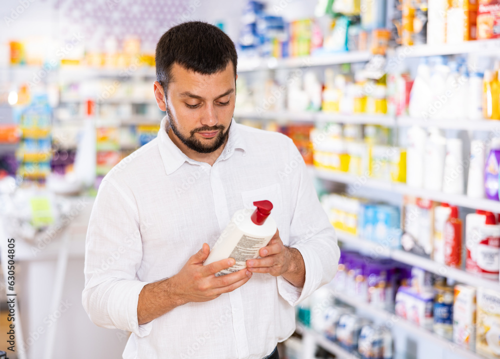 Positive young adult man customer choosing lotion or shampoo at drugstore