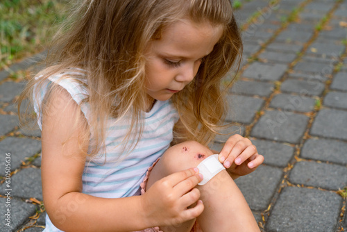 A little girl puts a band-aid on a wound on her knee after an accident. Selective focus.