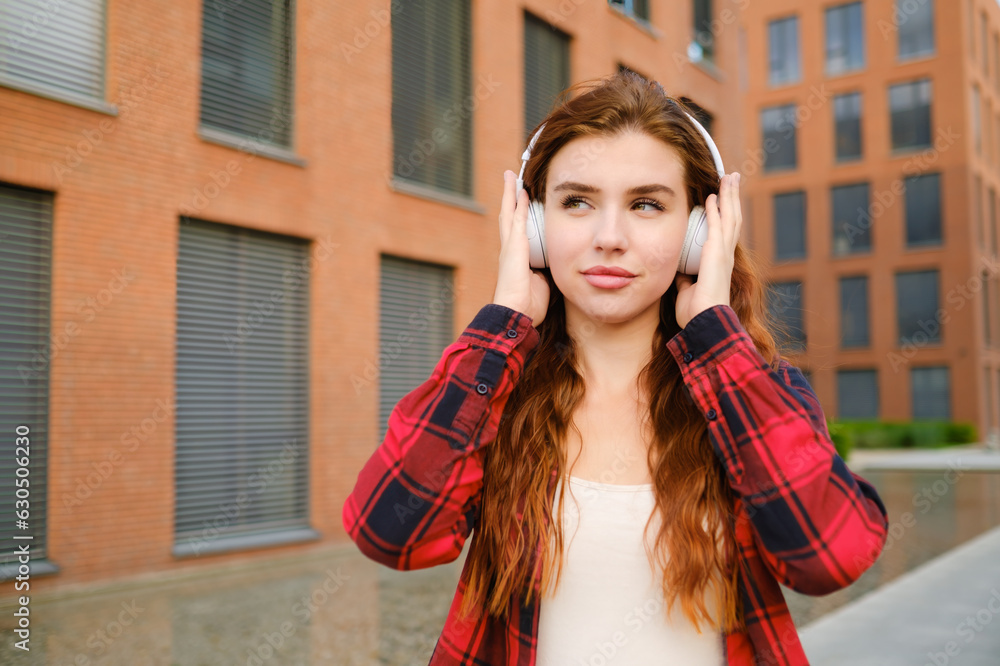 Young happy woman enjoying listening to music from headphones in the urban city center, wearing a checkered shirt. 