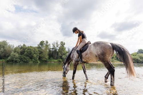 Girl riding gray horse down the calm river water with forest greenery reflections from the background. Nature and animal love concept.