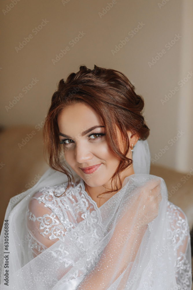 The morning of the bride in a white robe and an elegant hairstyle. An adult girl sits on a chair and fastens an earring, looking into the camera lens and smiling The morning of the bride in a white robe and an elegant hairstyle. An adult girl sits on a chair and fastens an earring, looking into the camera lens and smiling