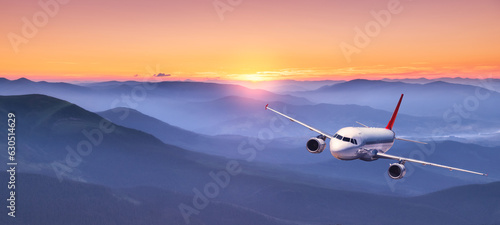 Plane is flying in colorful sky at sunset. Landscape with passenger airplane over mountains in fog, orange sky at twilight. Aircraft is landing. Business. Aerial view. Transport. Private Jet