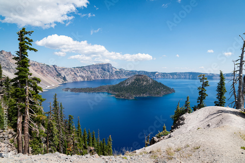 Horizontal vista with pine tree forest, rocks and island at Crater Lake on a sunny day with mostly blue sky. The park is located at Crater Lake National Park in Oregon. 