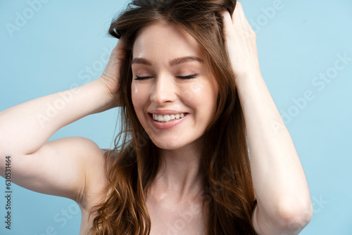 Smiling young woman taking shower, holding hair, doing head massage isolated on blue background