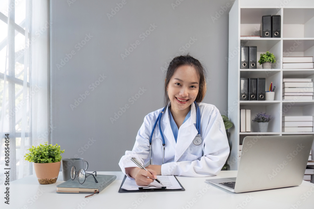 Asian doctor young beautiful woman smiling using working with a laptop computer and her writing something on paperwork or clipboard white paper at hospital desk office, Healthcare medical concept Asian doctor young beautiful woman smiling using working with a laptop computer and her writing something on paperwork or clipboard white paper at hospital desk office, Healthcare medical concept