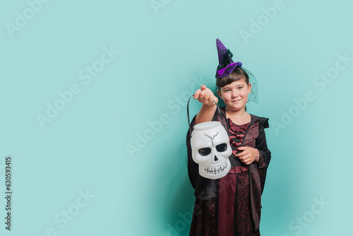 Happy laughing little girl in a carnival witch costume holding basket for treats on a color background