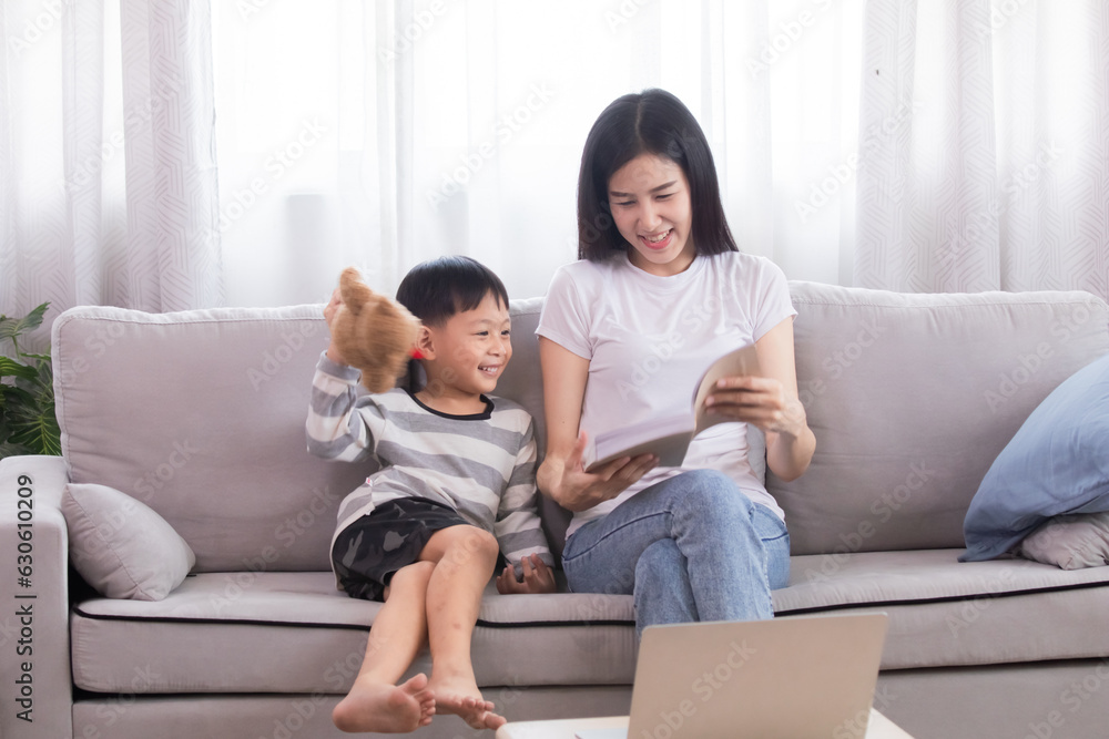Asian beautiful mother and son read favorite book together on weekends, young woman and adorable boy lying down on sofa reading a comic book in living room, Enjoy listening to stories from mom. Asian beautiful mother and son read favorite book together on weekends, young woman and adorable boy lying down on sofa reading a comic book in living room, Enjoy listening to stories from mom.