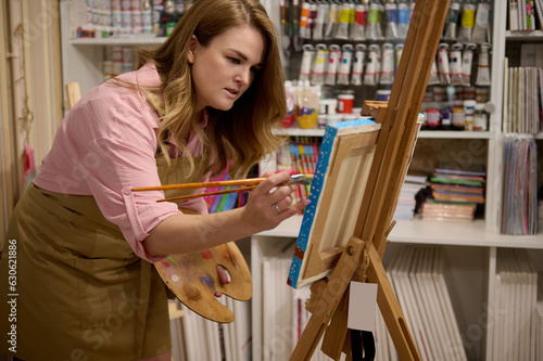 Inspired female painter draws on canvas, standing at easel in a creative art studio against a shelf with oil paint tubes