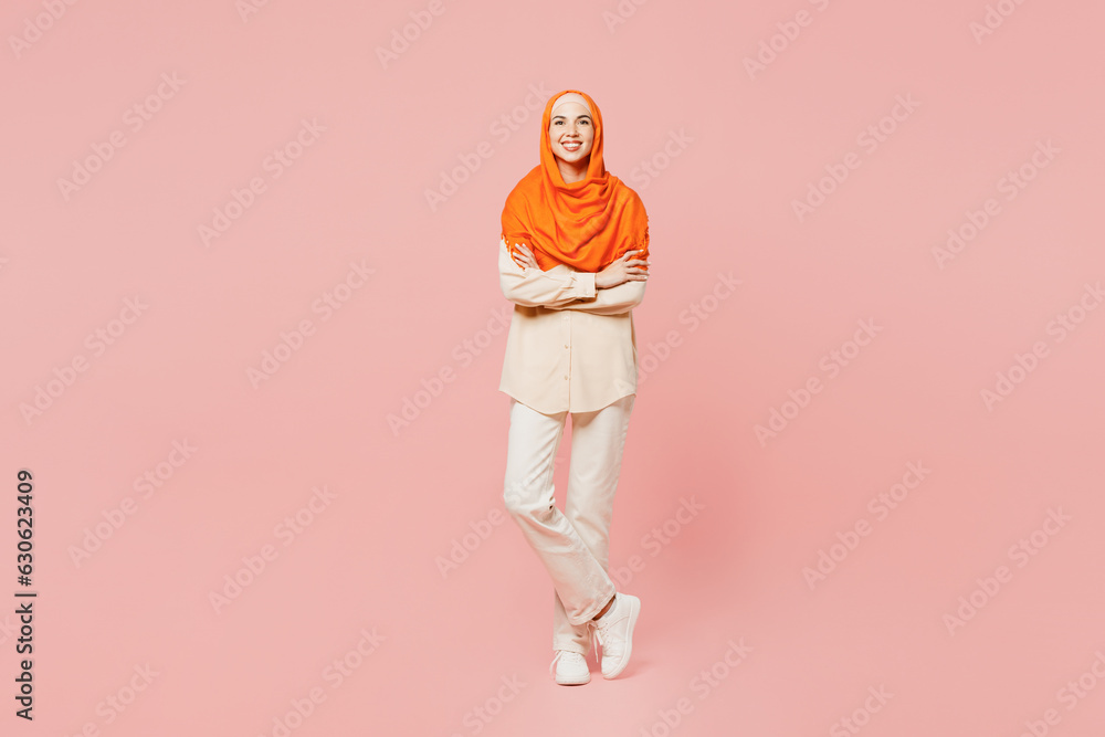 Fototapeta premium Full body smiling happy young arabian asian muslim woman wears orange abaya hijab hold hands crossed folded look camera isolated on plain pink background. Uae middle eastern islam religious concept.