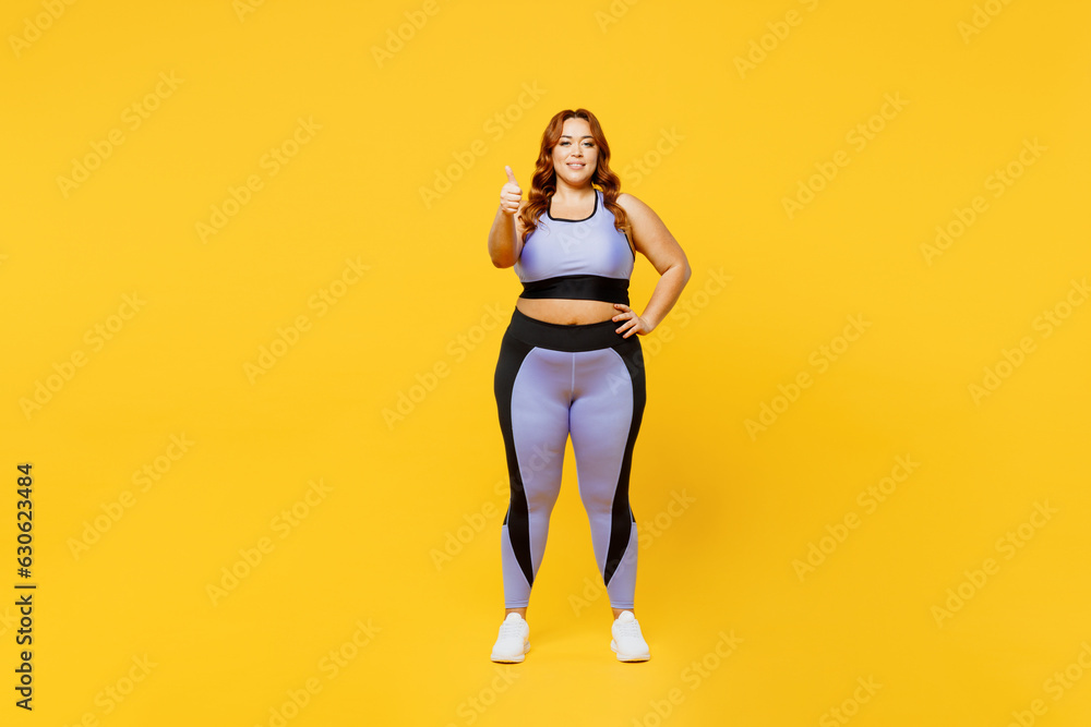 Full body smiling happy young chubby plus size big fat fit woman wears blue top warm up training look camera show thumb up isolated on plain yellow background studio home gym. Workout sport concept. Full body smiling happy young chubby plus size big fat fit woman wears blue top warm up training look camera show thumb up isolated on plain yellow background studio home gym. Workout sport concept.
