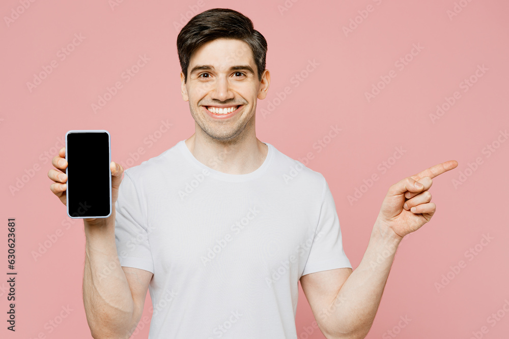 Young happy man wear white t-shirt casual clothes hold in hand using mobile cell phone in blue case with blank screen workspace area point aside isolated on plain pastel light pink background studio. Young happy man wear white t-shirt casual clothes hold in hand using mobile cell phone in blue case with blank screen workspace area point aside isolated on plain pastel light pink background studio.