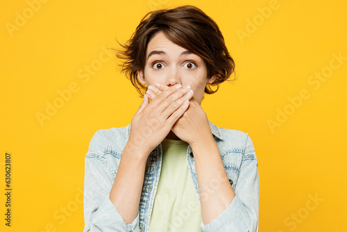 Young shocked sad scared woman she wears green t-shirt denim shirt casual clotheslook camera cover mouth with hand look camera isolated on plain yellow background studio portrait. Lifestyle concept.