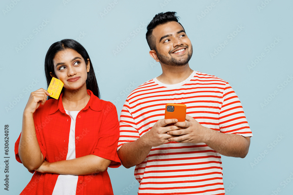Young minded couple two friend family Indian man woman in red casual clothes t-shirt use mobile cell phone credit bank card shopping online book tour together isolated on plain blue color background