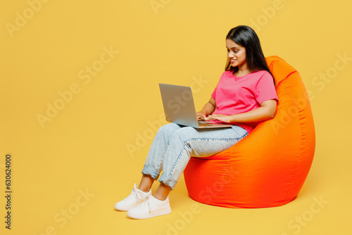 Full body young happy IT Indian woman wearing pink t-shirt casual clothes sit in bag chair hold use work on laptop pc computer isolated on plain yellow background studio portrait. Lifestyle concept.