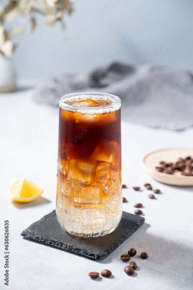 Coffee with tonic in a tall glass with ice and lemon on a light blue background with coffee beans and fruit. Trendy summer craft refreshing drink. Coffee with tonic in a tall glass with ice and lemon on a light blue background with coffee beans and fruit. Trendy summer craft refreshing drink.