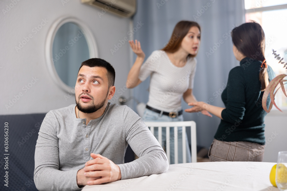 Bored man sitting at table while two female relatives arguing behind