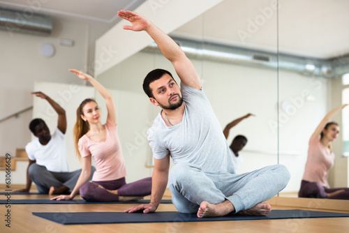 Young bearded man doing stretching exercises during group training in fitness center  sitting in lotus position  bending sideways with arm raised overhead