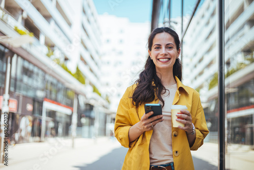 Modern young woman walking on the city street texting and holding cup of coffee. Business woman holding smartphone and looking away outdoors. 