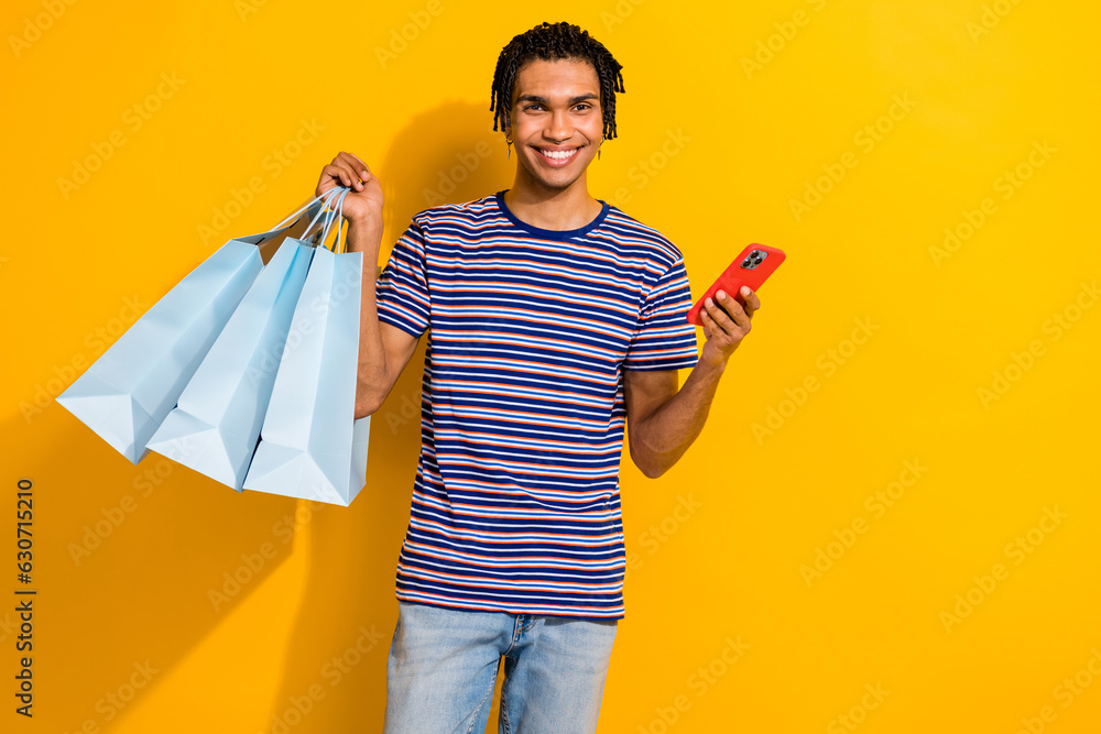 Portrait of toothy beaming man with piercing wear stylish t-shirt hold shopping bags smartphone isolated on yellow color background Portrait of toothy beaming man with piercing wear stylish t-shirt hold shopping bags smartphone isolated on yellow color background