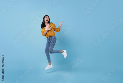 Cheerful positive Asian girl jumping in the air with pointing finger to copy space with smile face and happy isolated on blue background.