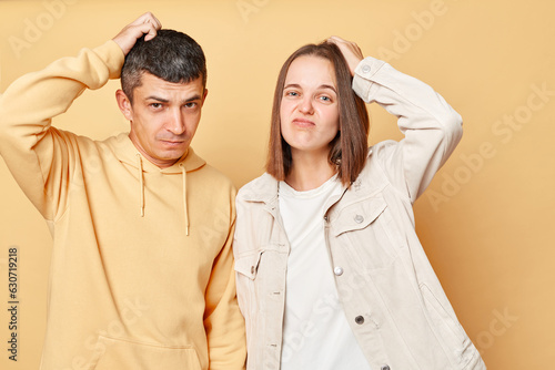 Konstfotografi Confused puzzled young man and woman couple standing together isolated over beige background standing rubbing head looking at camera with doubtful face