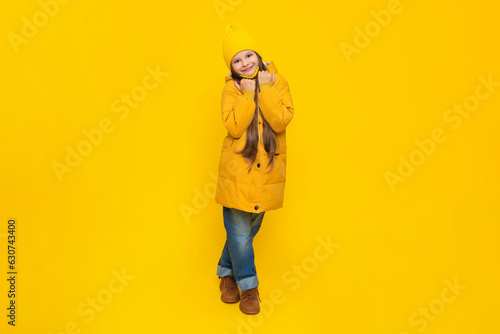 A young girl rejoices at the onset of autumn. Warm clothes for children. A child in an orange down jacket and hat on a yellow isolated background in the studio.