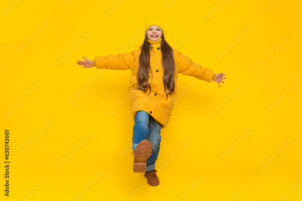 A little girl in full height in an autumn coat, down jackets and hats, rejoices in autumn. The child joyfully raises his leg with his arms outstretched to the sides. Yellow isolated background.