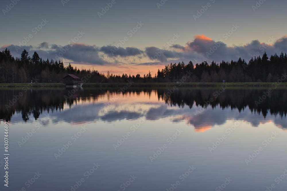 Fototapeta premium Tranquil landscape of a lake surrounded by lush landscape, with the sun setting in the background