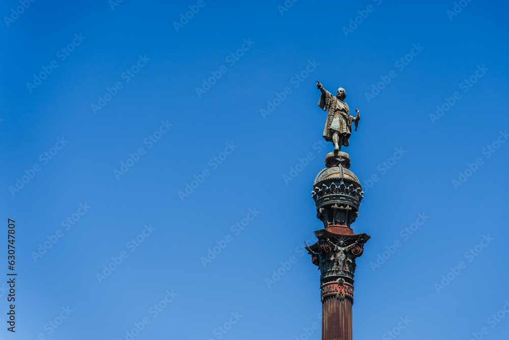 Monument and clock tower stand tall against a bright blue sky: Columbus monument in Barcelona