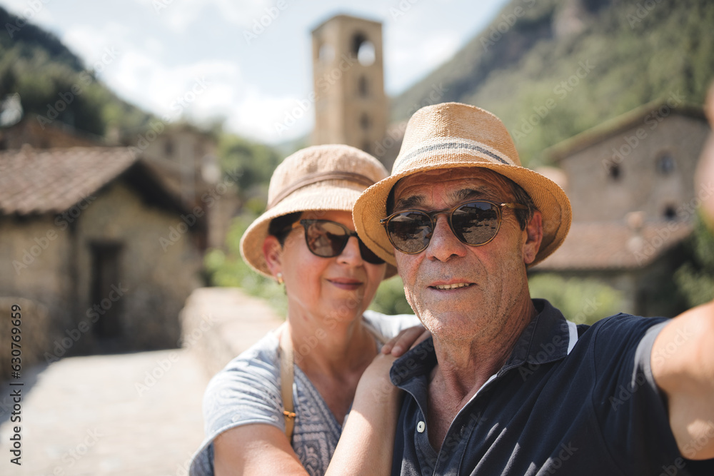 Older couple taking selfie with smartphone outside - Two elderly tourists enjoying summer vacation together in Beget village, Girona, Catalonia - Lifestyle concept with mature woman and man smiling at