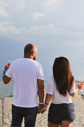  Portrait of a happy Smiling beautiful young couple in while standing at the beach.Happy casual couple holding hands and hugging and walking at the beach holding glasses of wine