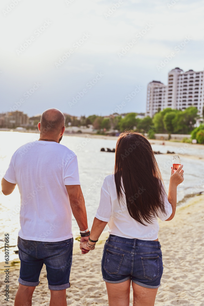 Portrait of a happy Smiling beautiful young couple in while standing at the beach.Happy casual couple holding hands and hugging and walking at the beach  holding glasses of wine
