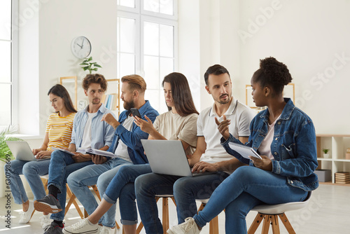 Diverse people sitting on chairs in row holding electronic devices. Candidates in casual clothes holding phones, laptop and resumes preparing for job interview in office. Human resources concept