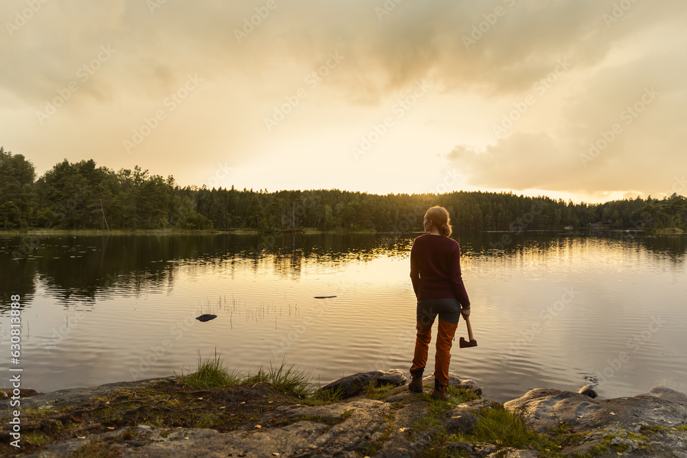 A female hiker holding an axe standing in front of a calm lake in the forest watching the sunset.